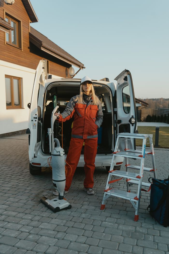 who-we-are A cleaner in uniform stands beside a van with cleaning tools outside a modern house at sunset.